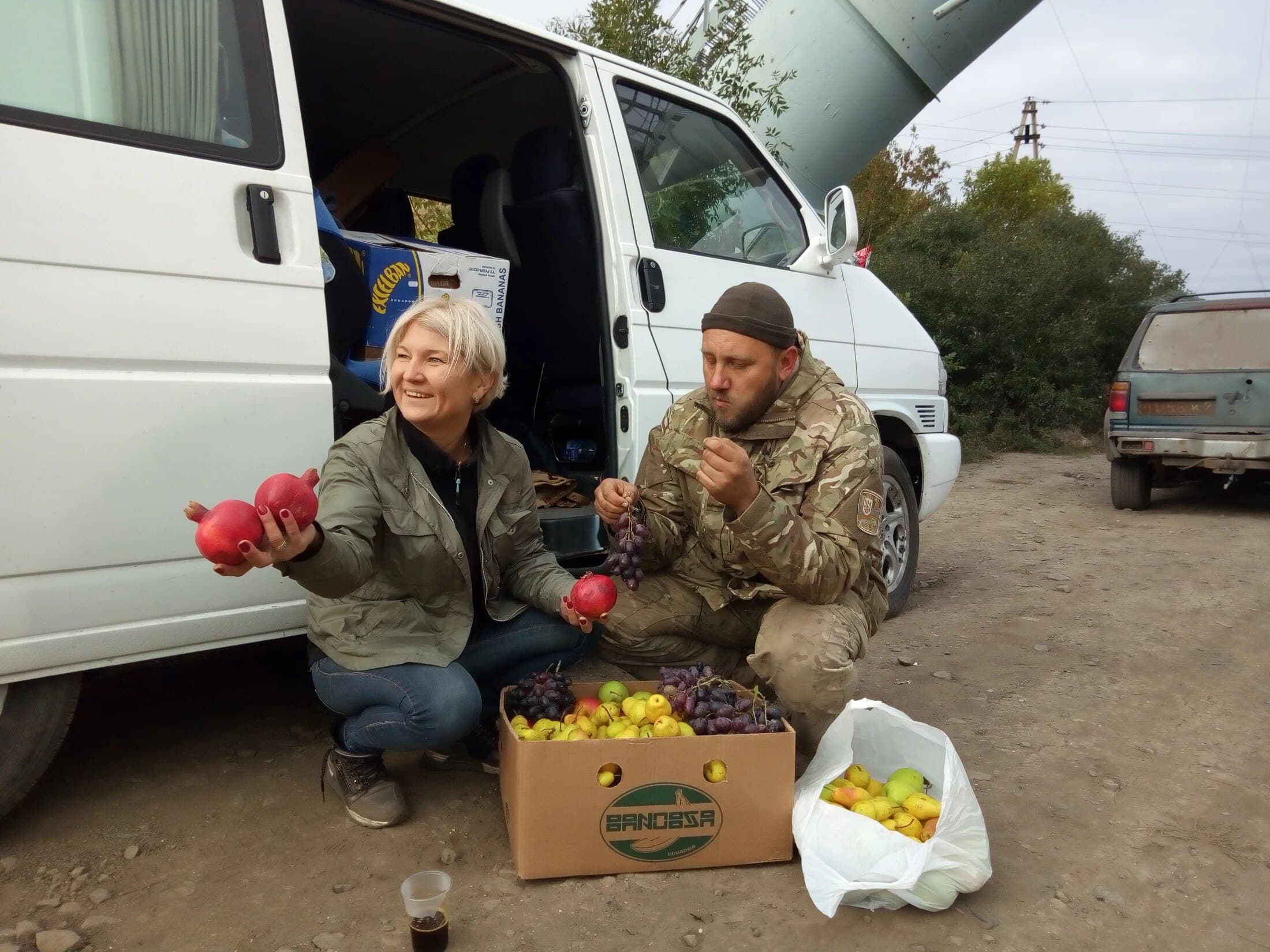 Natalka laughing beside the aid van, holding out pomegranates over a box of fresh fruit
